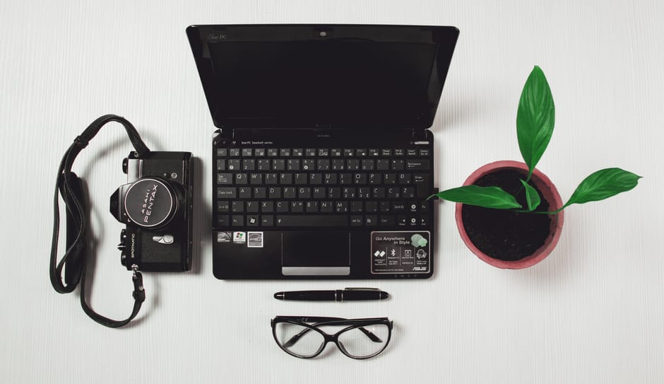 Top view of a workspace with laptop, camera, potted plant, pen, and glasses on a white surface.