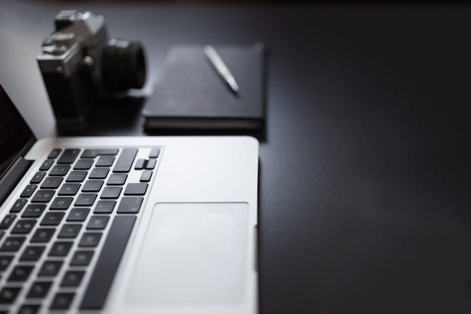 Modern workspace featuring a laptop, camera, notebook, and pen on a sleek black desk.